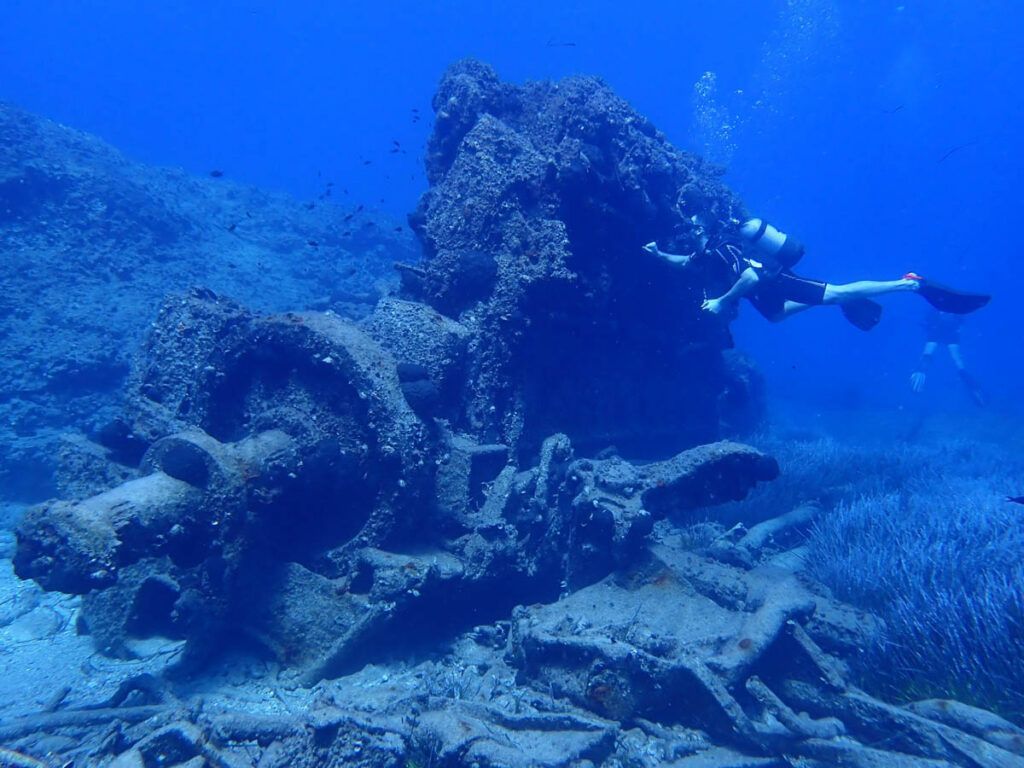 Scuba diver swimming alongside large marine machinery and twisted metal wreckage