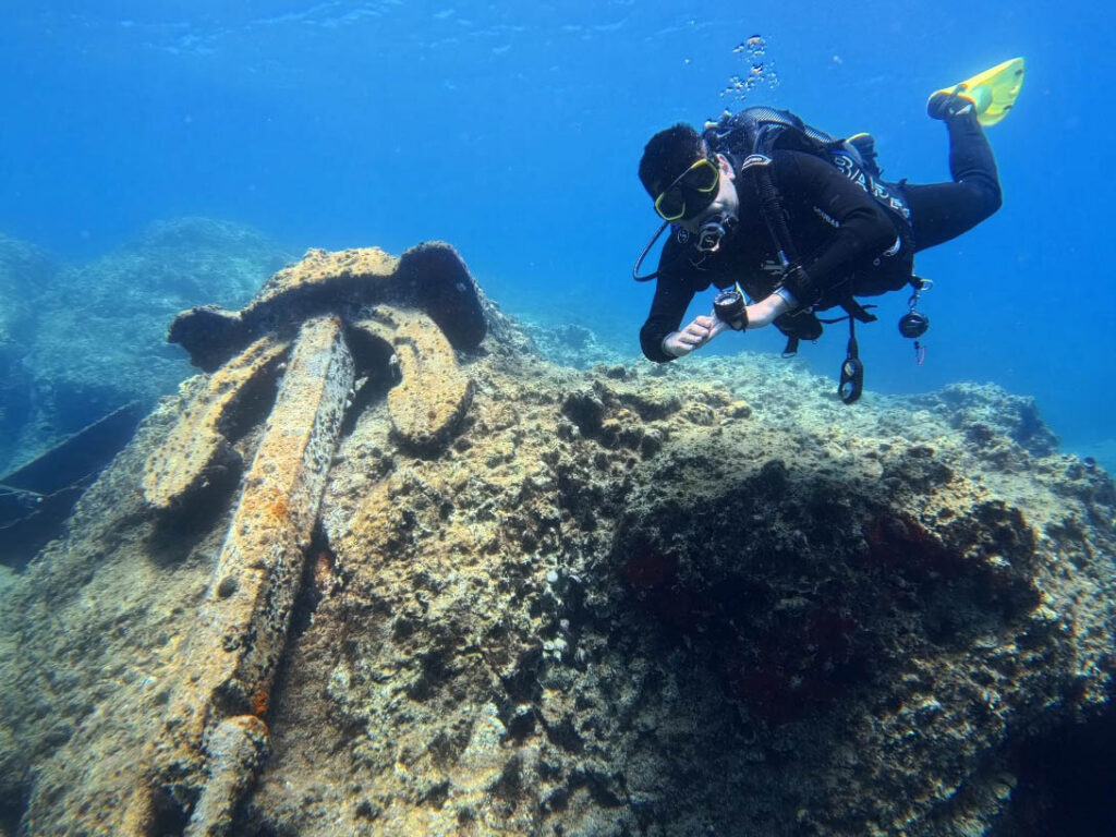 Scuba diver hovering beside a large encrusted anchor among rocks at the Karpaz wreck site